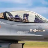 Un cadru apropiat de tip profil al cabinei unui avion de vânătoare F-16 în care se observă pilotul cu cască și echipament militar complet