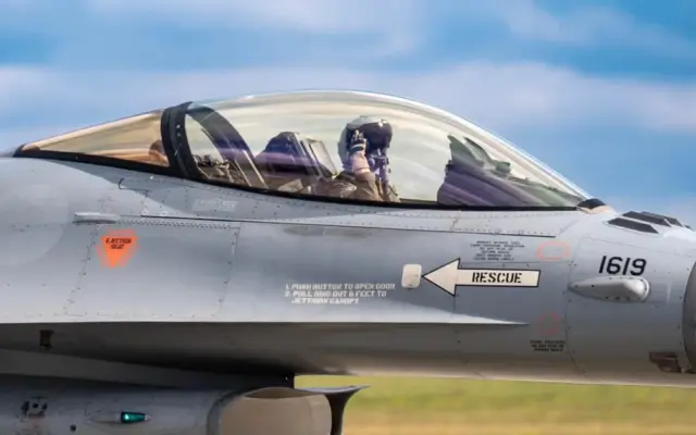 Un cadru apropiat de tip profil al cabinei unui avion de vânătoare F-16 în care se observă pilotul cu cască și echipament militar complet
