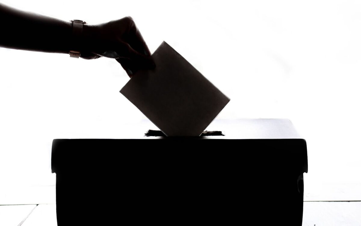 O siluetă a unei mâini care introduce un plic de vot într-o urnă, pe un fundal alb luminos
