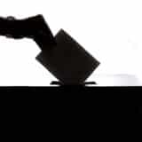 O siluetă a unei mâini care introduce un plic de vot într-o urnă, pe un fundal alb luminos