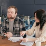 Un bărbat și o femeie stau la o masă de bucătărie și analizează niște documente care par a fi facturi