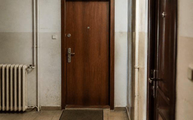 O imagine din interiorul unui bloc care prezintă ușa unui apartament, de culoare maro închis cu aspect de lemn, aflată la capătul unui coridor