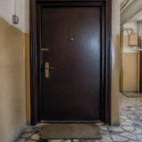O ușă de apartament de culoare maro închis, cu vizor, mâner auriu și un lanț de siguranță, situată pe un hol de bloc cu pereți bej și podea din mozaic
