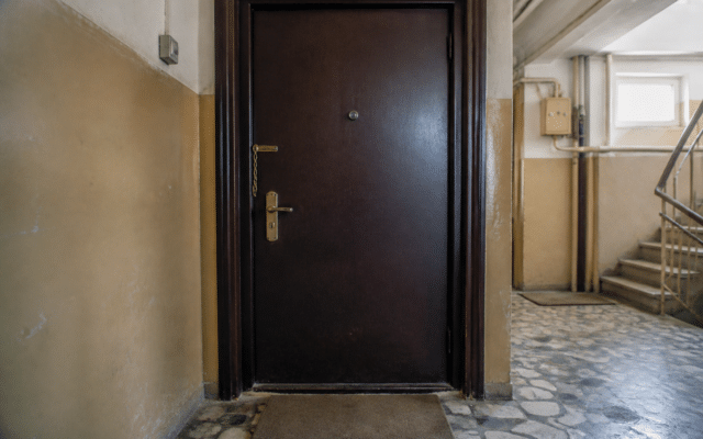 O ușă de apartament de culoare maro închis, cu vizor, mâner auriu și un lanț de siguranță, situată pe un hol de bloc cu pereți bej și podea din mozaic