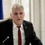 Un bărbat cu părul grizonant, purtând un costum negru, cămașă albă și cravată roșie, este așezat la o masă și vorbește