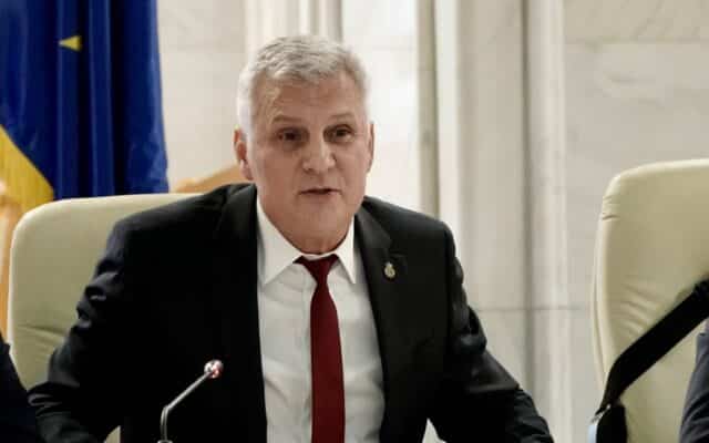 Un bărbat cu părul grizonant, purtând un costum negru, cămașă albă și cravată roșie, este așezat la o masă și vorbește