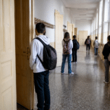 Un hol lung de școală, cu uși înalte din lemn pe partea stângă și ferestre pe partea dreaptă