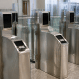 Un șir de porți automate de control la frontieră, realizate din metal și sticlă, echipate cu ecrane negre pentru recunoaștere facială și cititoare de documente