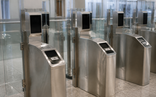 Un șir de porți automate de control la frontieră, realizate din metal și sticlă, echipate cu ecrane negre pentru recunoaștere facială și cititoare de documente
