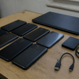 Mai multe telefoane mobile negre așezate pe o masă de lemn, toate cu ecranul în jos, lângă un laptop închis, un hard disk extern și două cabluri USB