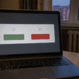 Un laptop deschis pe o masă, într-o cameră cu un calorifer și o fereastră spre niște blocuri la amurg