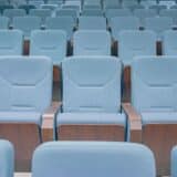 O fotografie de aproape a mai multor rânduri de scaune de cinema sau dintr-o sală de conferințe, de culoare albastru deschis, cu spătare și cotiere din lemn