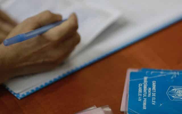 O mână ține un pix albastru și pare să scrie pe un document sau într-un catalog școlar, cu o adâncime de câmp mică