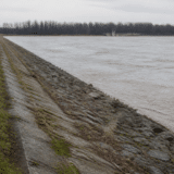 Un dig din beton și piatră consolidat pe malul unui râu mare cu apă tulbure, cu un drum pavat care se întinde în perspectivă