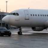 Un avion alb de pasageri parcat pe o pistă de aeroport, iar în fața lui se află o mașină de poliție de culoare albastru închis cu girofaruri
