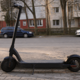 O trotinetă electrică neagră parcată pe trotuar, lângă bordura unei străzi